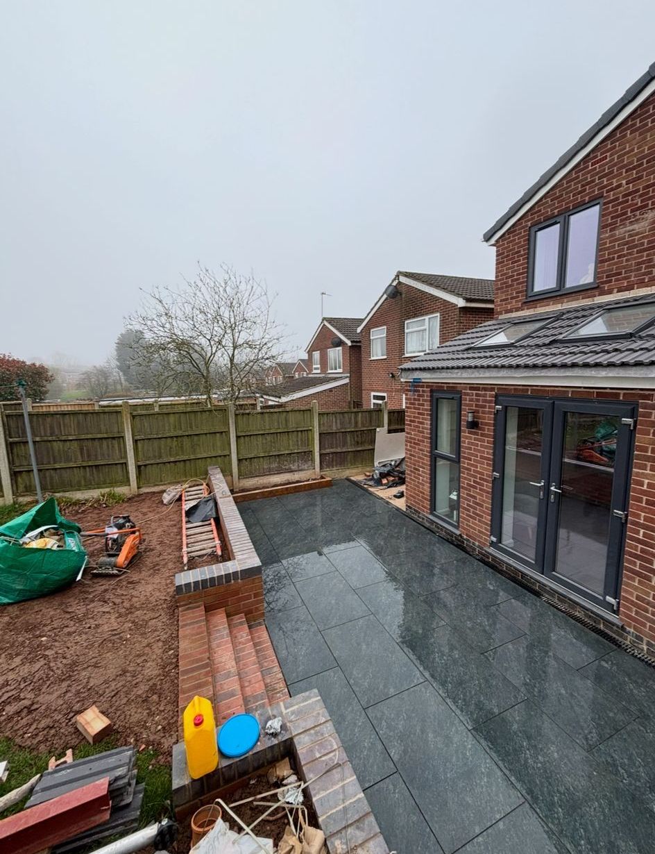 Backyard patio with gray paving, red-brick house, fenced lawn, and garden tools on an overcast day