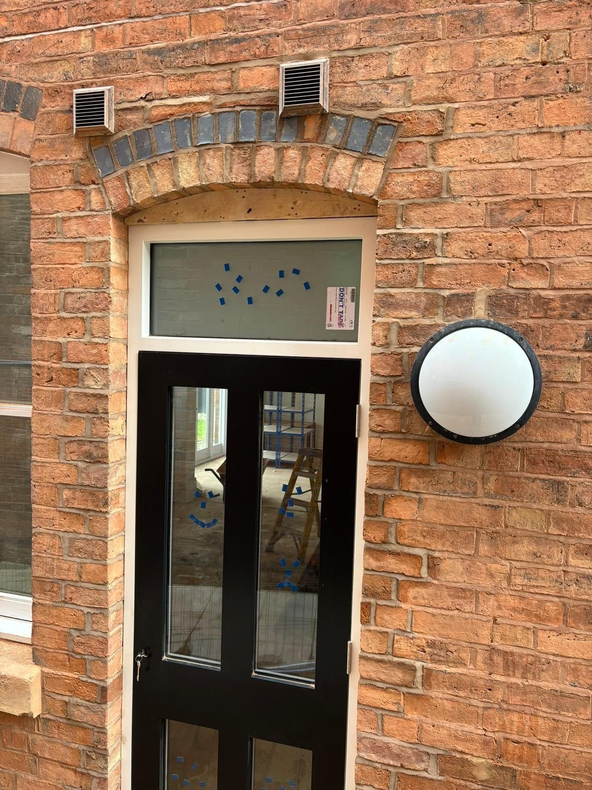 Brick building entrance with black door, arched brickwork, and round wall light above the doorway
