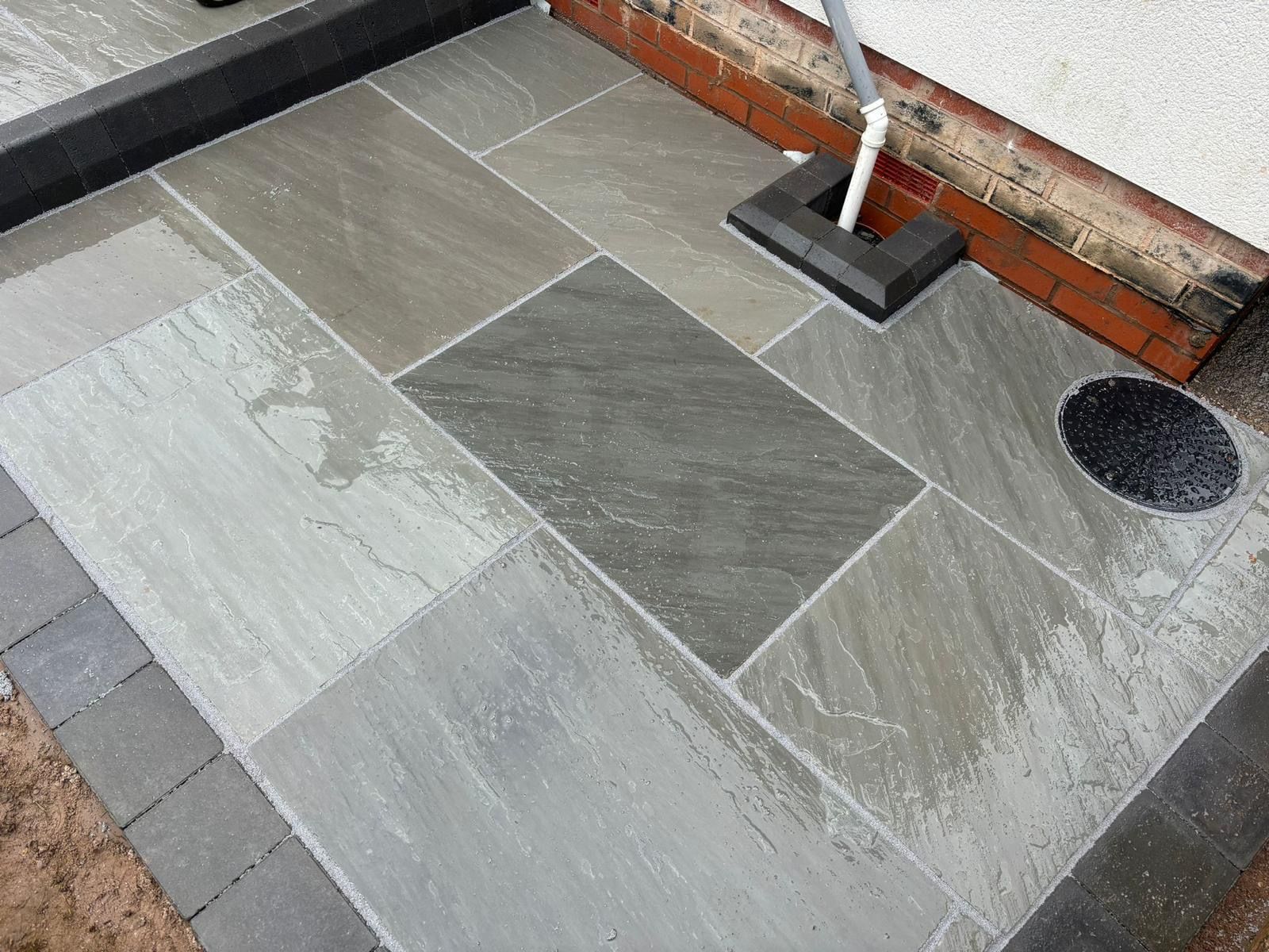 Outdoor tiled patio floor with gray stone tiles, a drain, and a broom handle near a brick wall