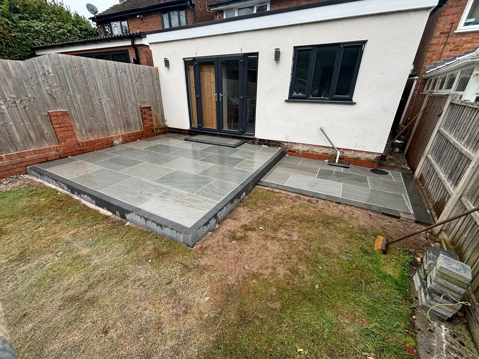 Backyard patio with gray paving slabs, white house wall, and a grassy lawn in front.