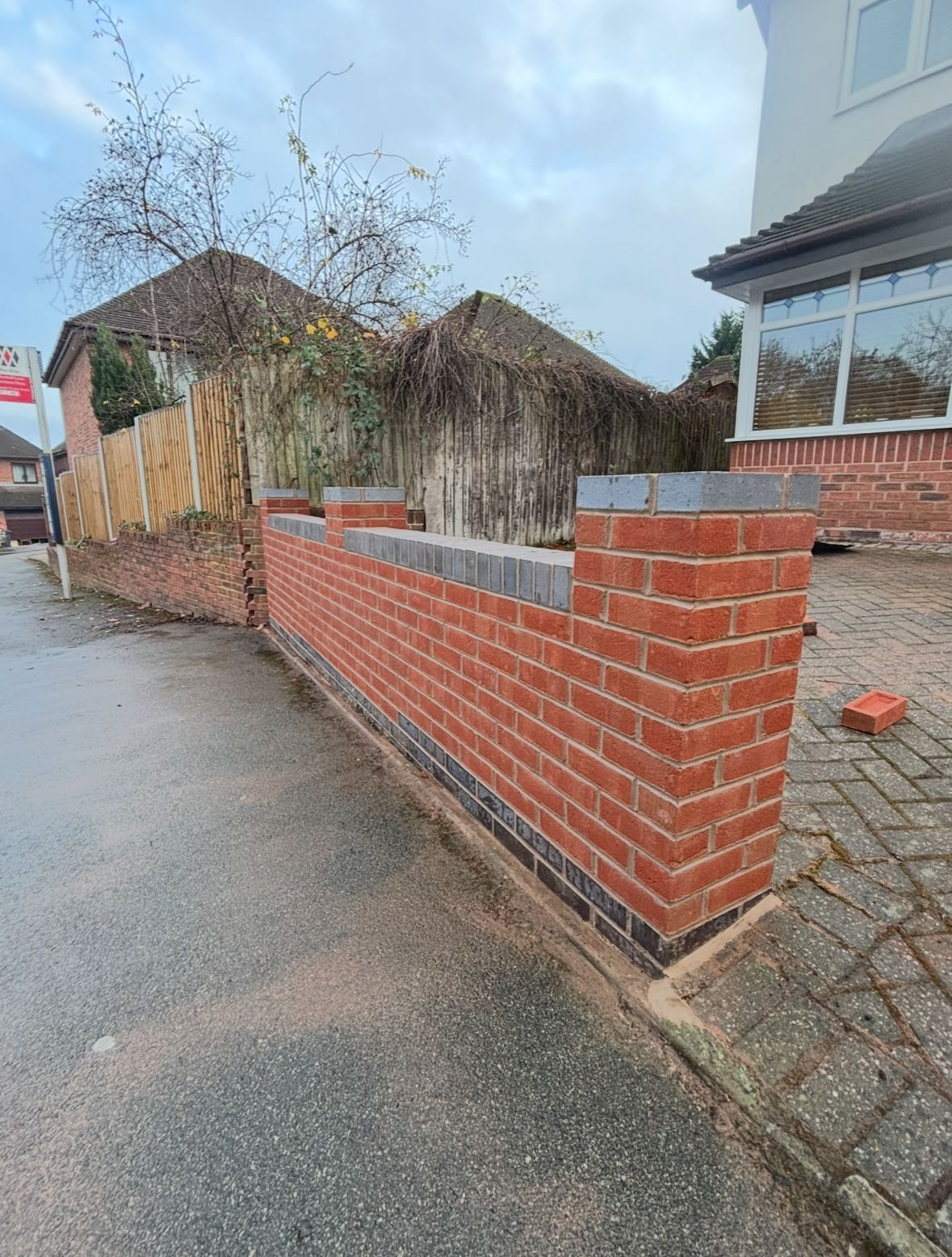 Wet street beside a red brick wall and house, with leafless shrubs and a small pile of bricks.