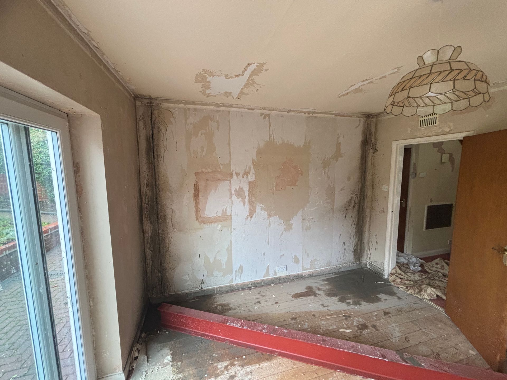 Damp, damaged room with peeling walls, stained ceiling, debris on the floor, and a red plank across the entrance