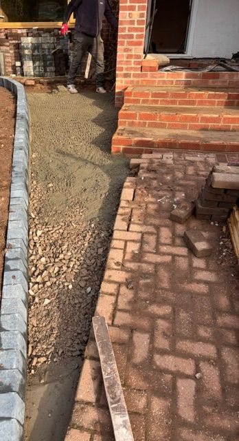 Brick walkway beside a house under construction, with gravel and a narrow dirt path along the foundation