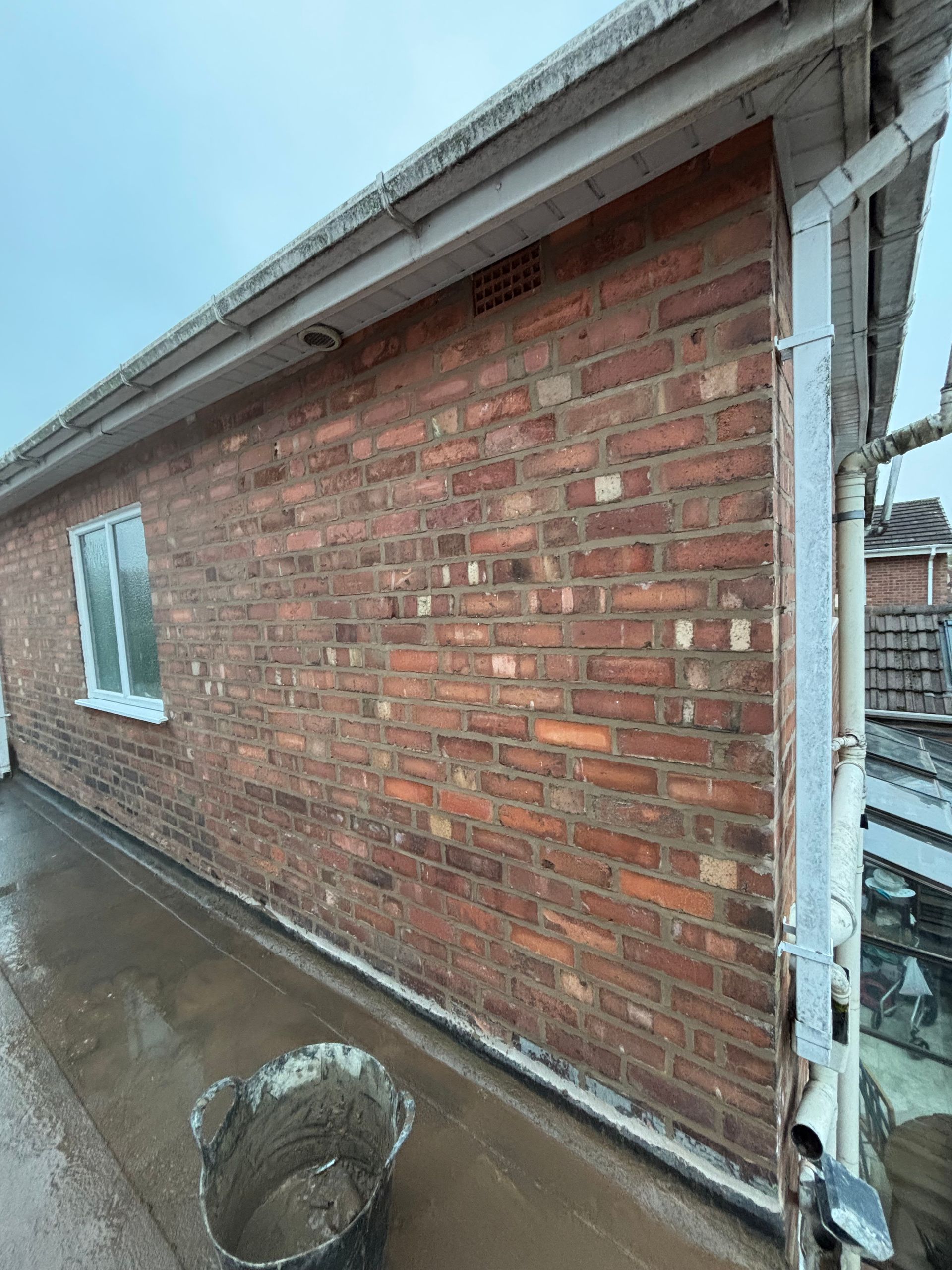 Brick house side wall with white trim and wet concrete walkway beside a bucket.