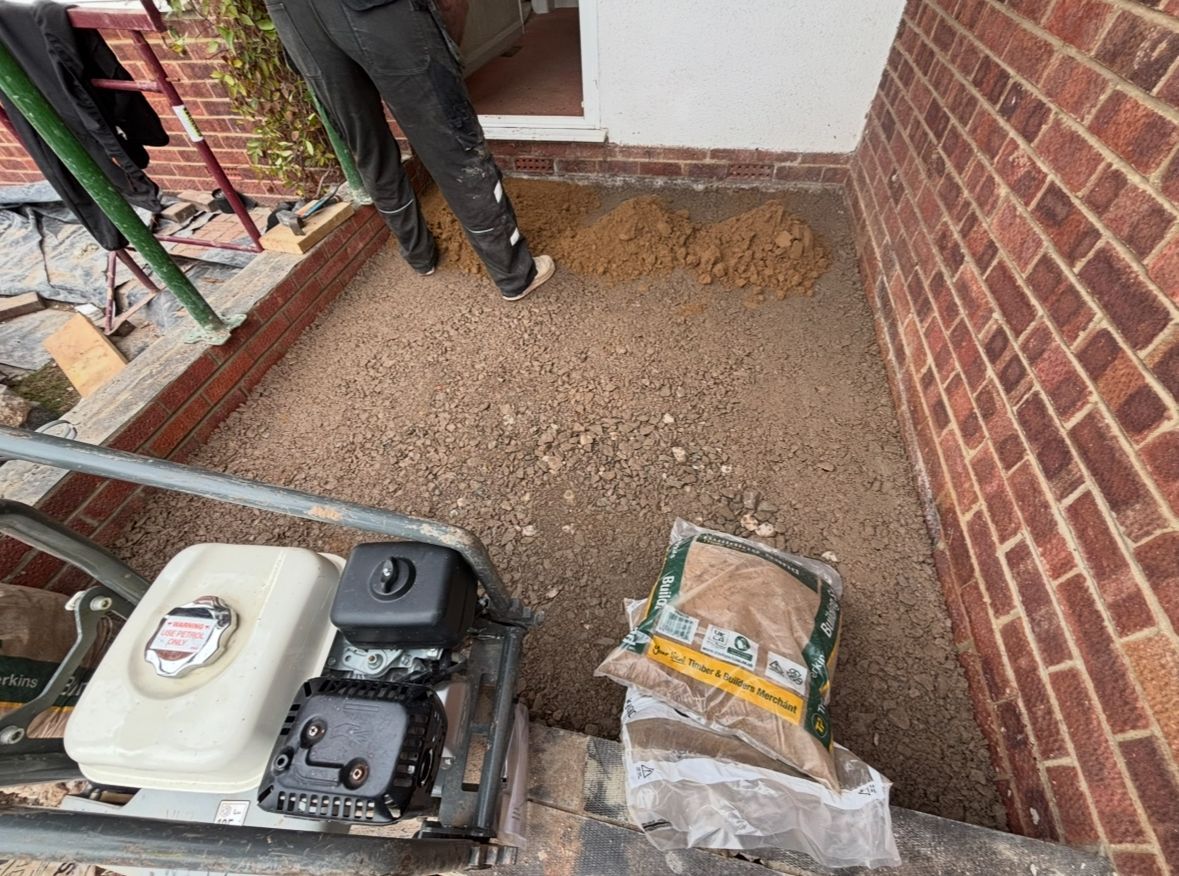 Workers leveling gravel in a small brick-lined patio beside a pressure washer and bag of material