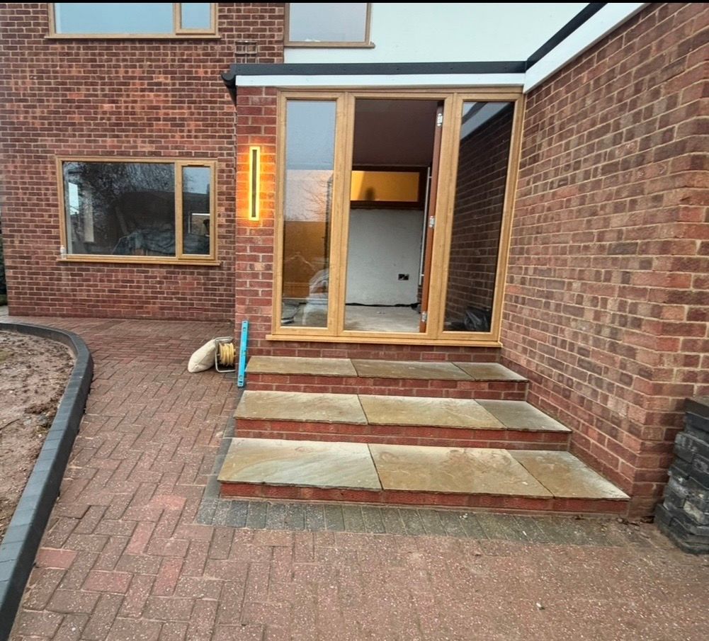 Brick patio leading to open wooden doors at a house entrance with steps and a lit wall sconce