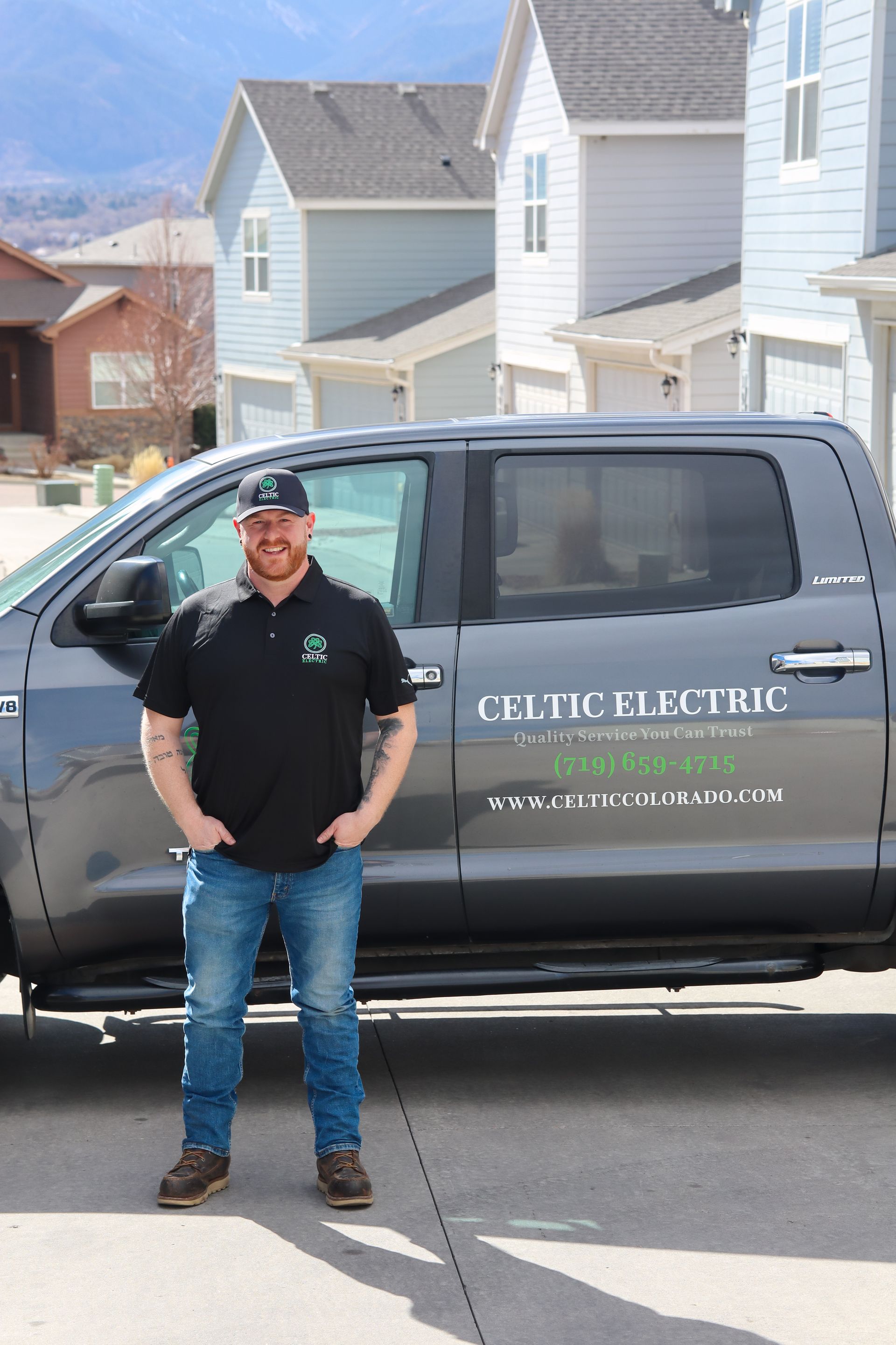 Man in black shirt with logo standing by gray truck. Background has houses and mountains.