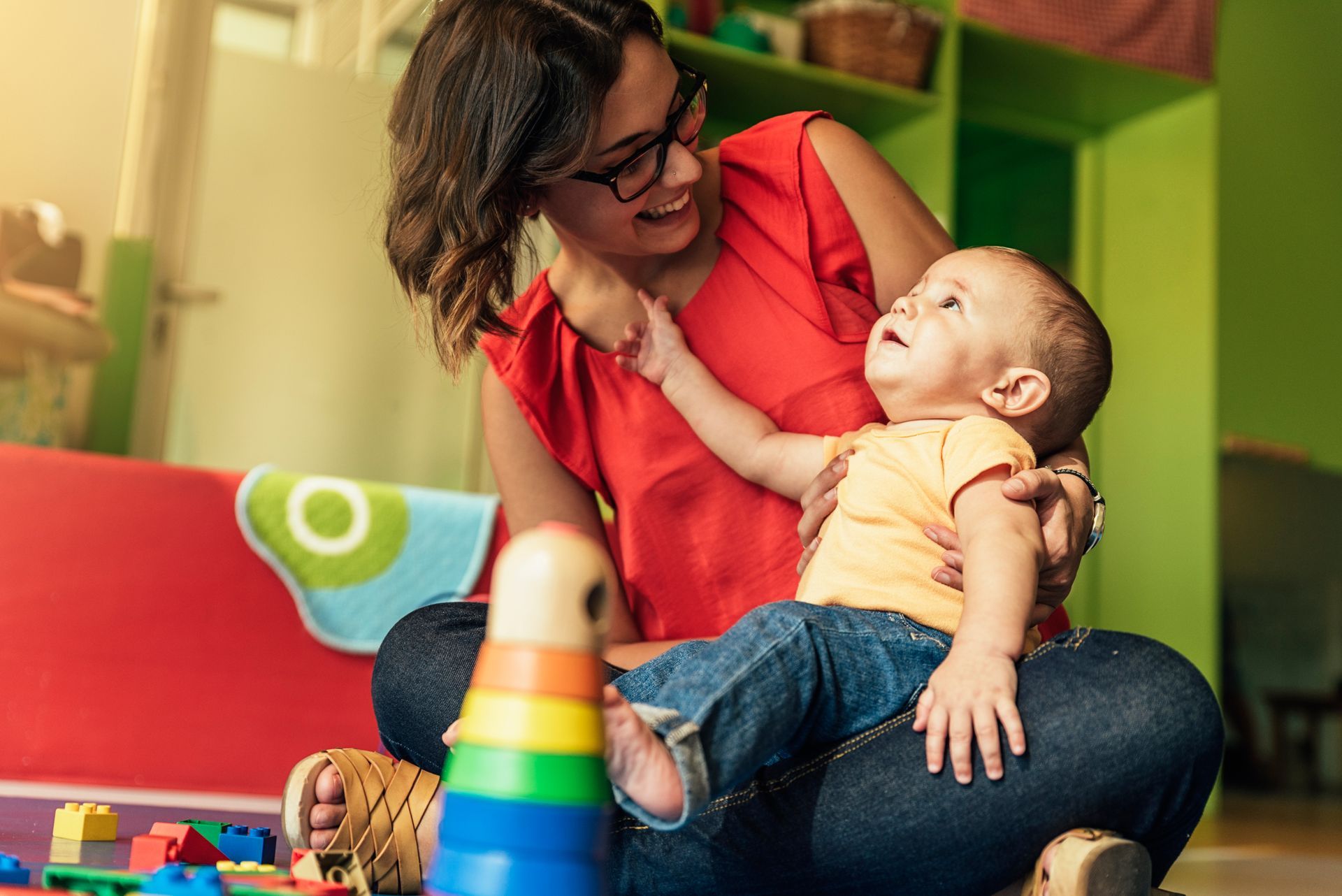 Smiling adult in red top holding a baby in a colorful playroom with toys