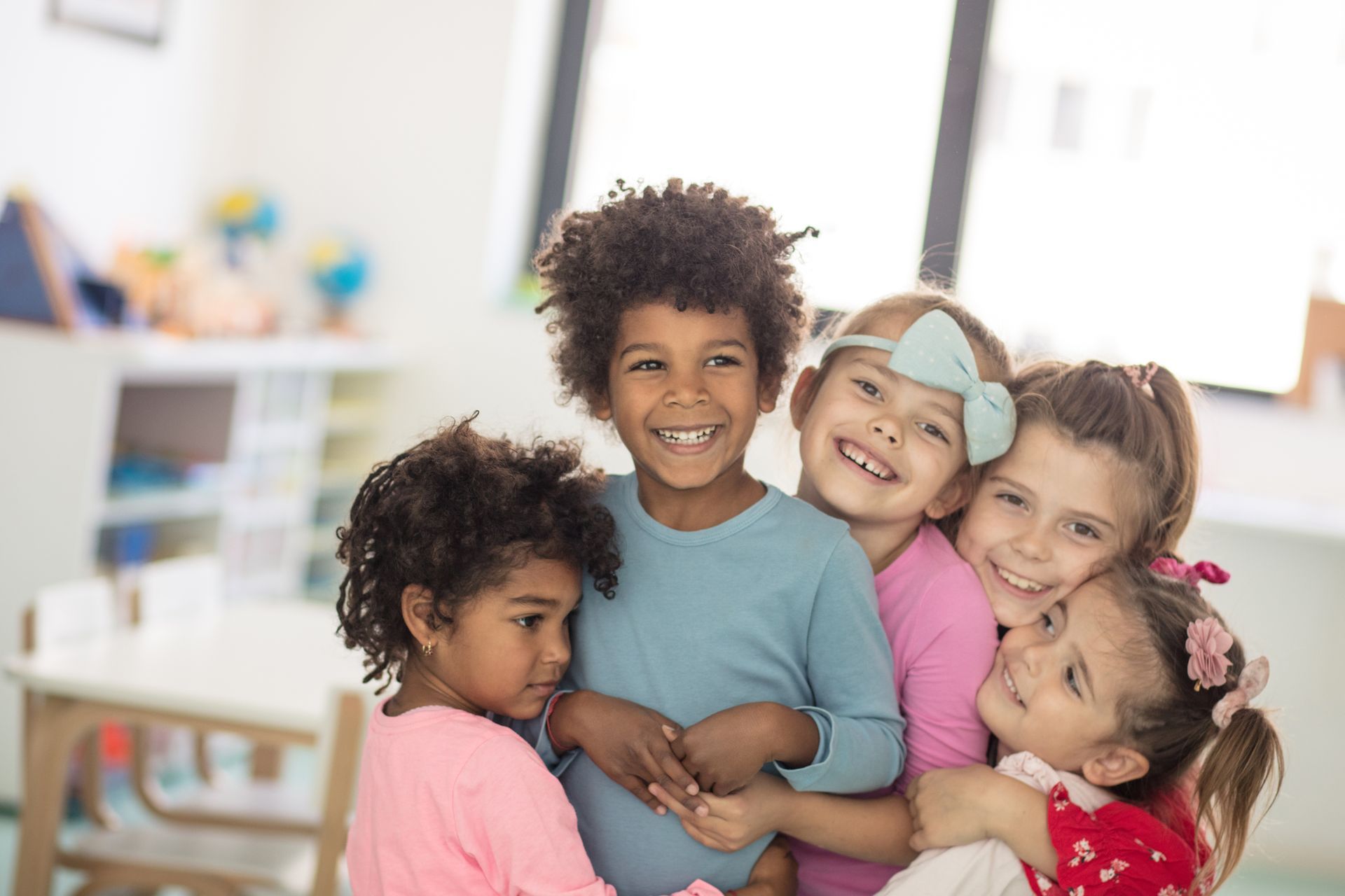 Five smiling children hugging together in a bright classroom or playroom.