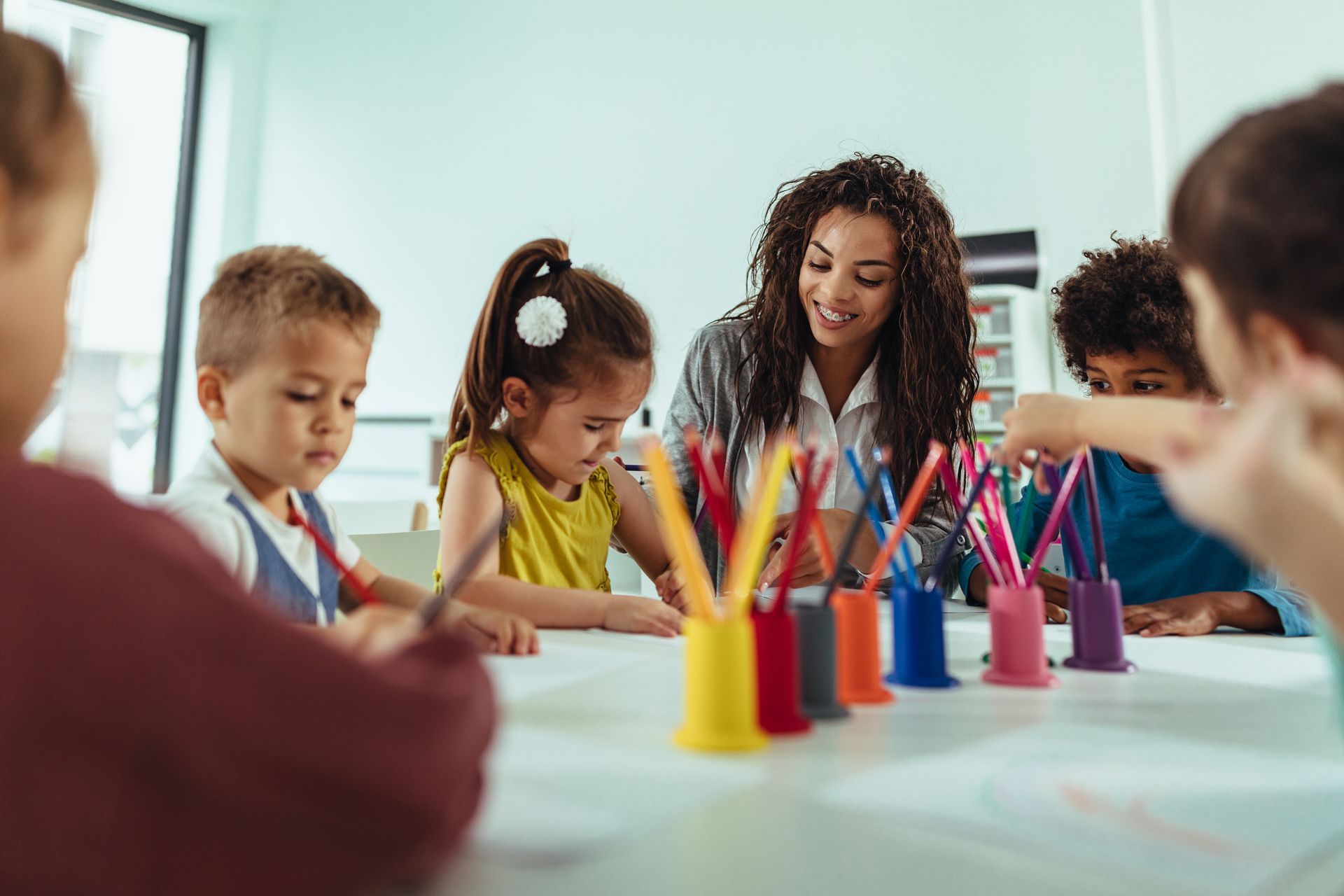 Children painting at a table with colorful art supplies while a teacher helps them in a classroom