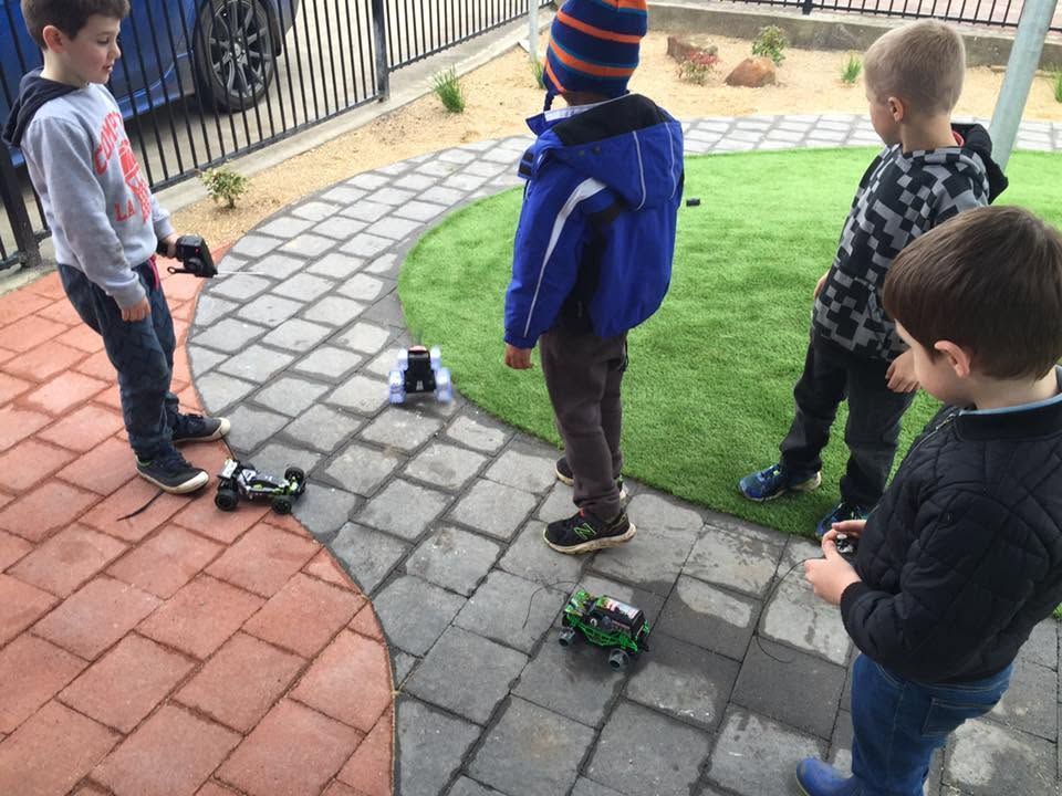 Children standing with small toy cars on a paved play area beside a grassy patch