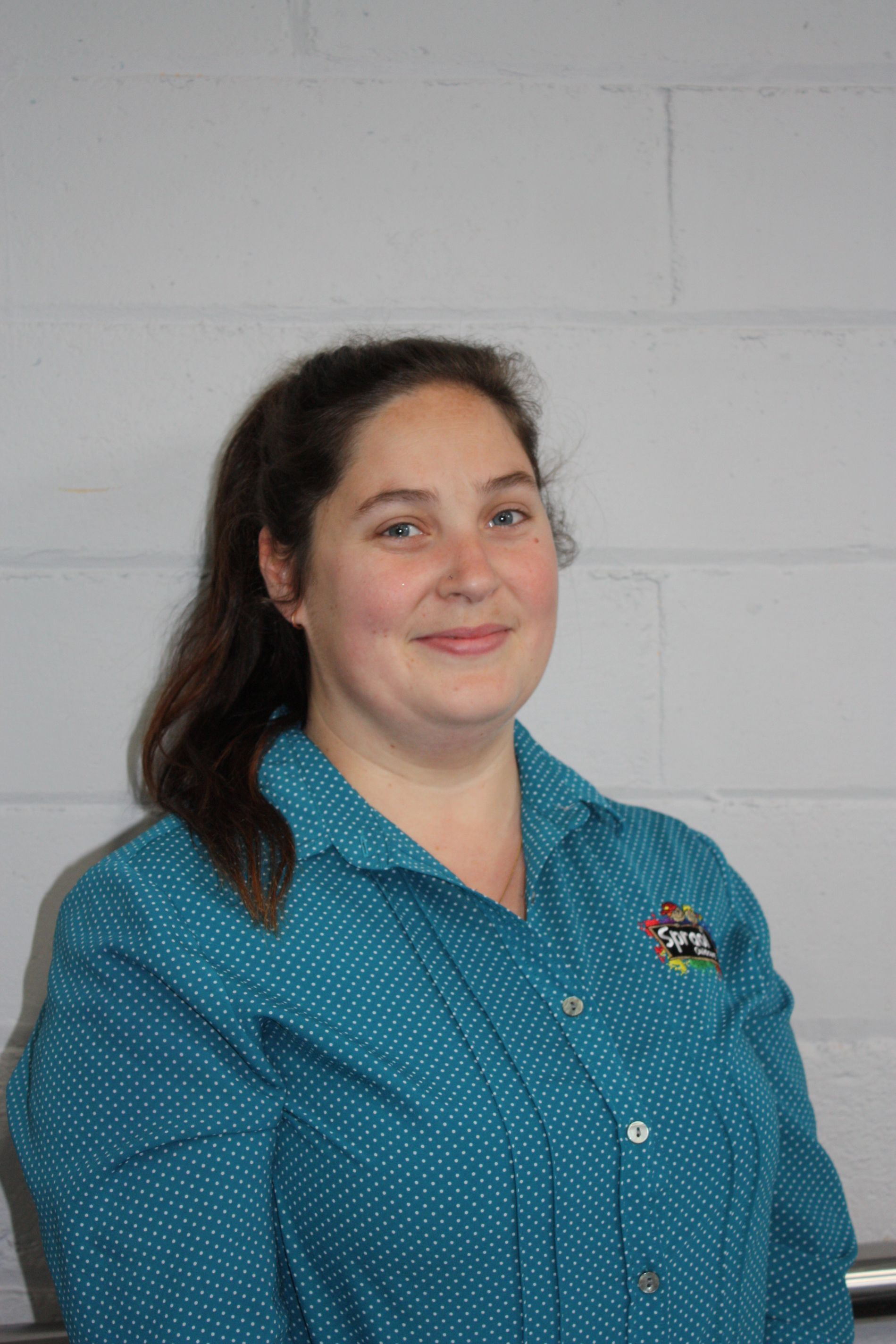 Woman in a teal shirt standing against a light gray brick wall, smiling at the camera.