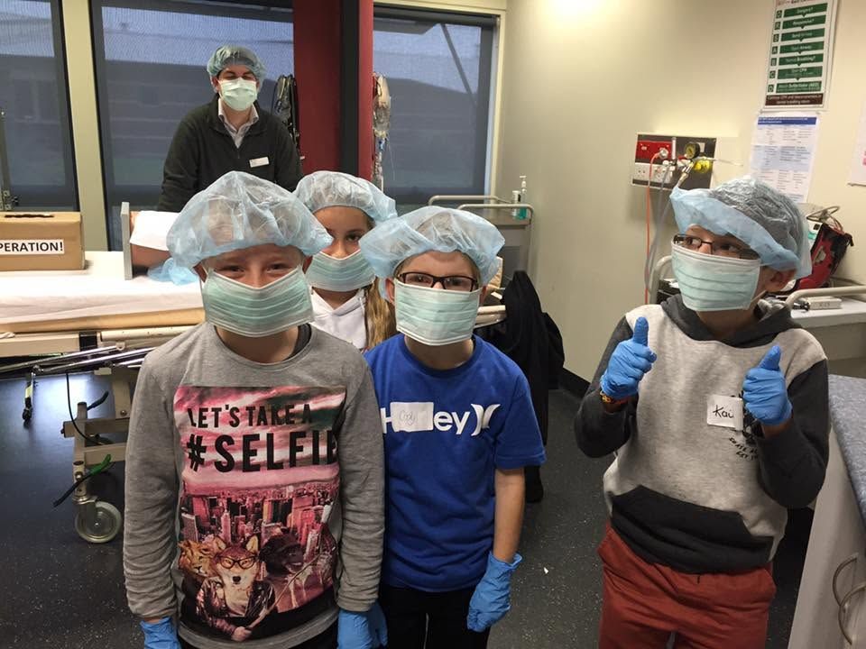 Four masked children in blue hairnets and gloves stand in a classroom or lab with a masked adult behind them.