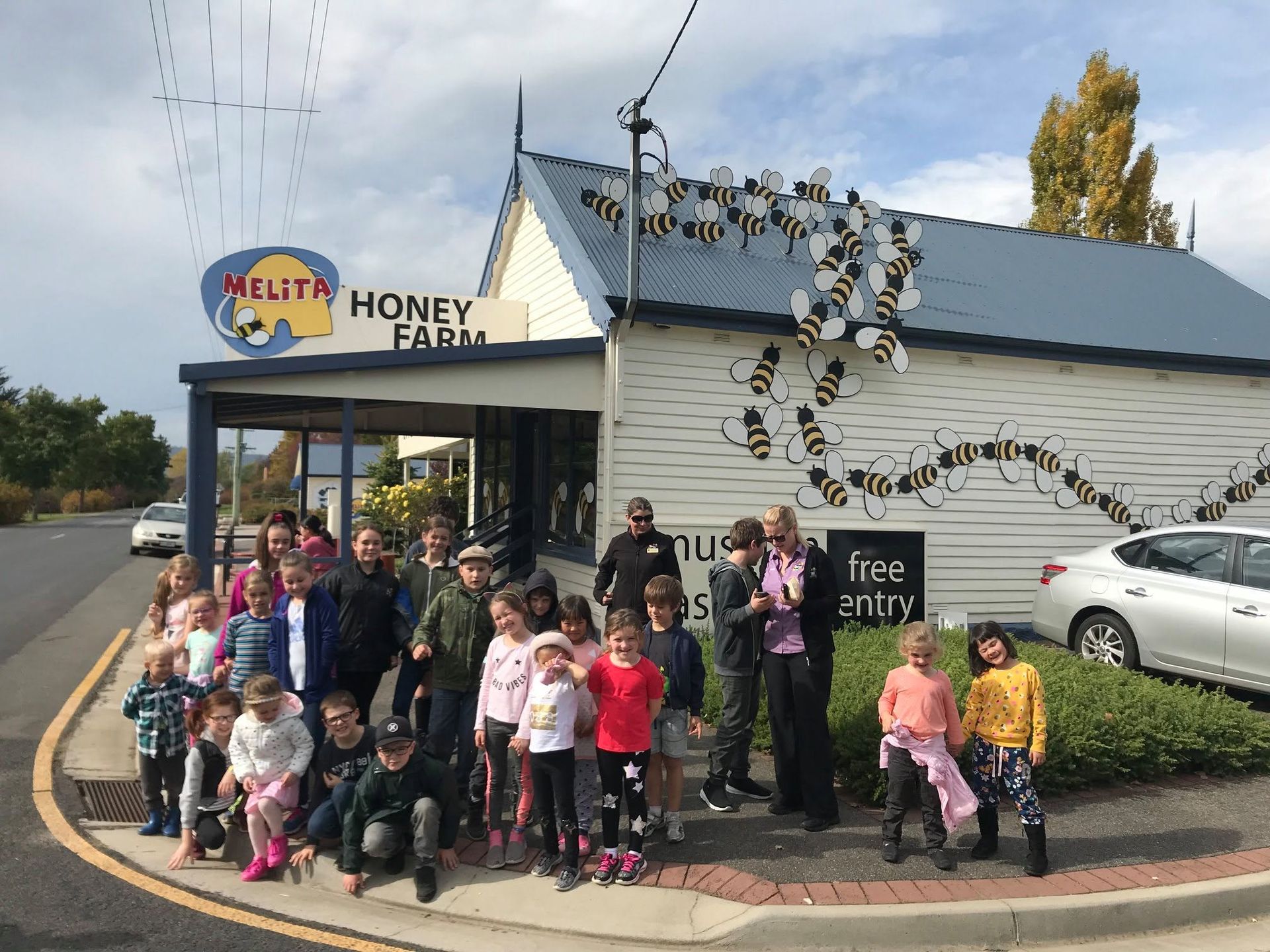 Group of people posing outside a honey shop with a bee sculpture and parked cars nearby