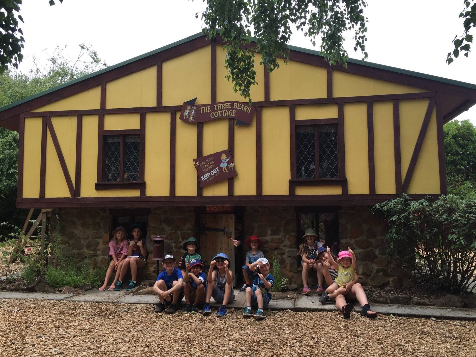 Children sitting outside a yellow-and-brown timber lodge in a wooded setting