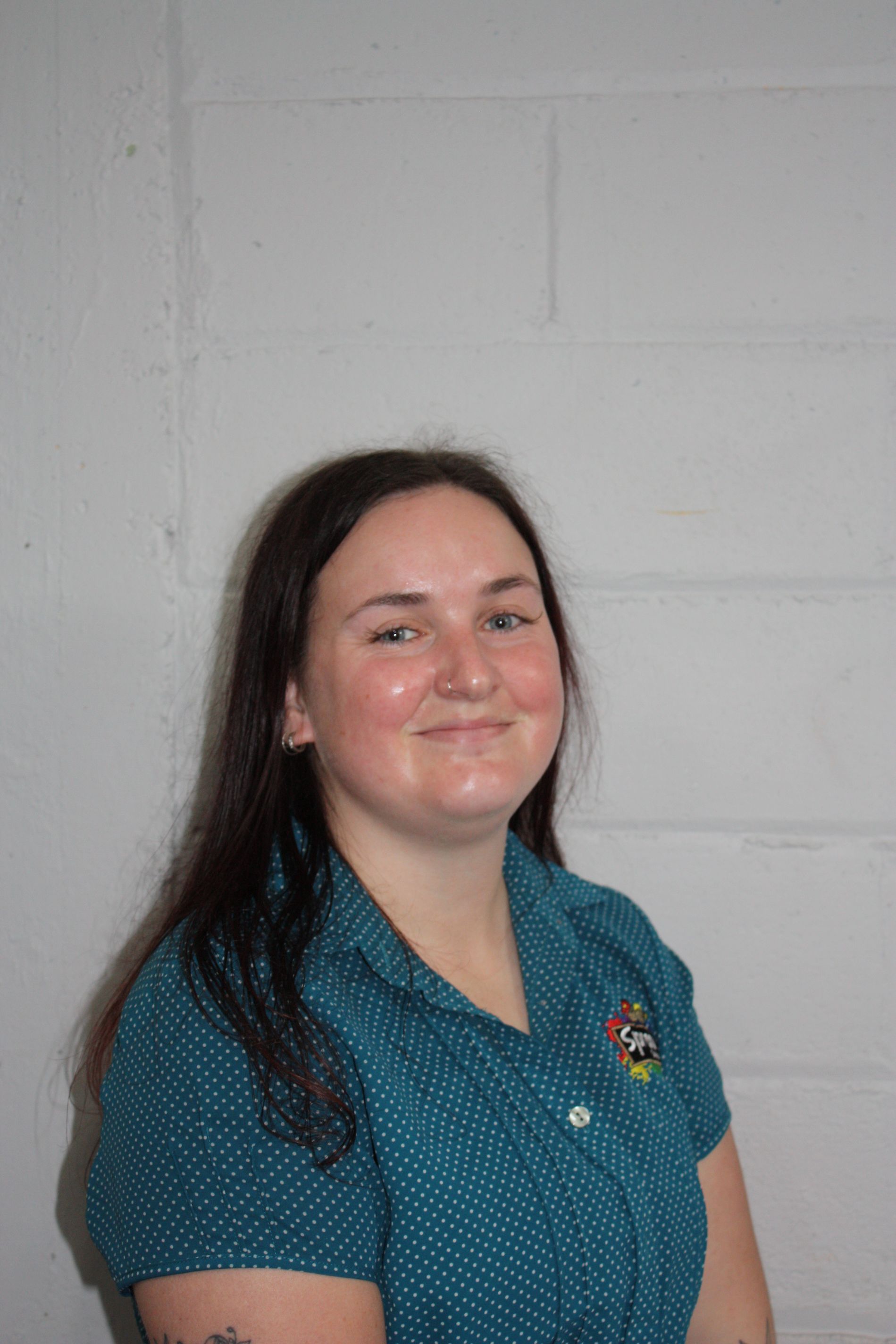 Woman in a teal shirt standing against a light gray wall, smiling at the camera