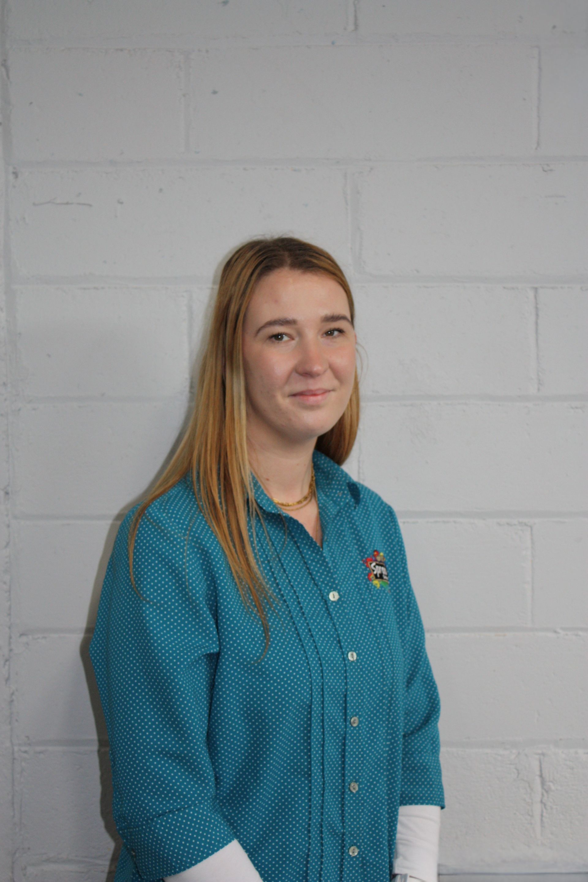 Woman in a teal polka-dot shirt standing against a light gray brick wall