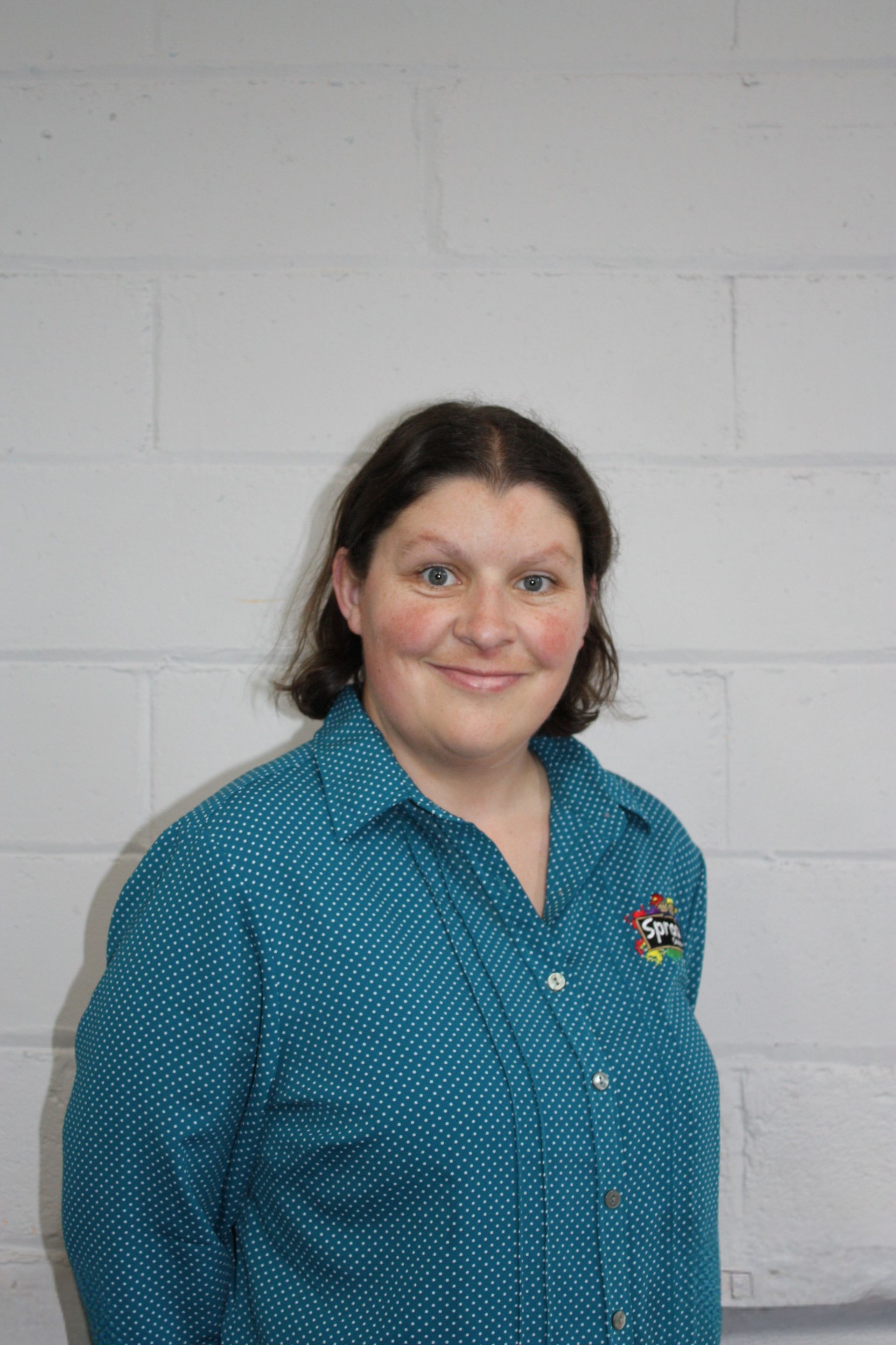 Woman in a teal polka-dot shirt standing against a white brick wall, smiling at the camera.