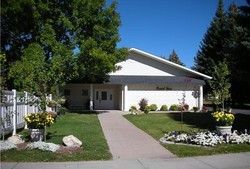 White building with angled roof, sidewalk, and green lawn. Trees and flowers in front.