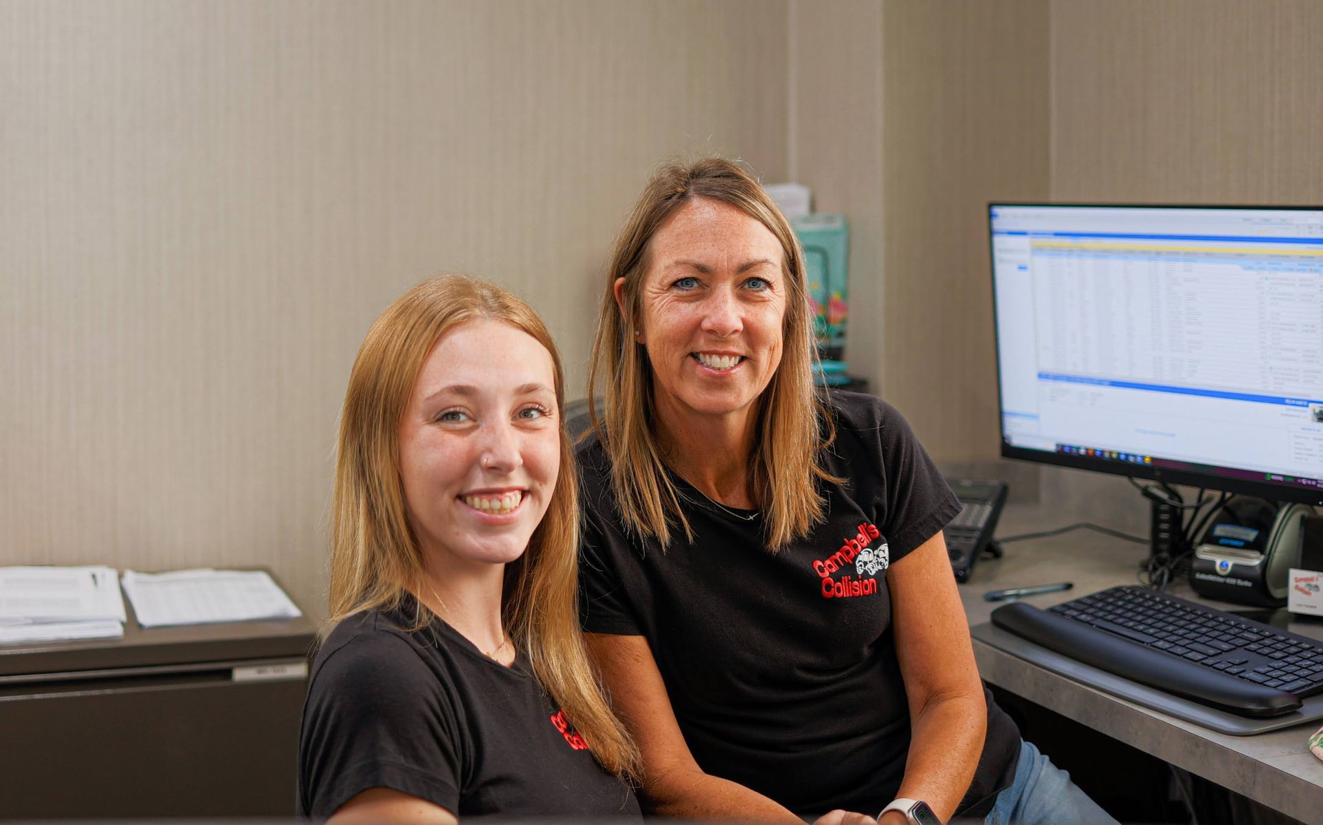 Two coworkers in black shirts smiling at a desk with a computer monitor in an office