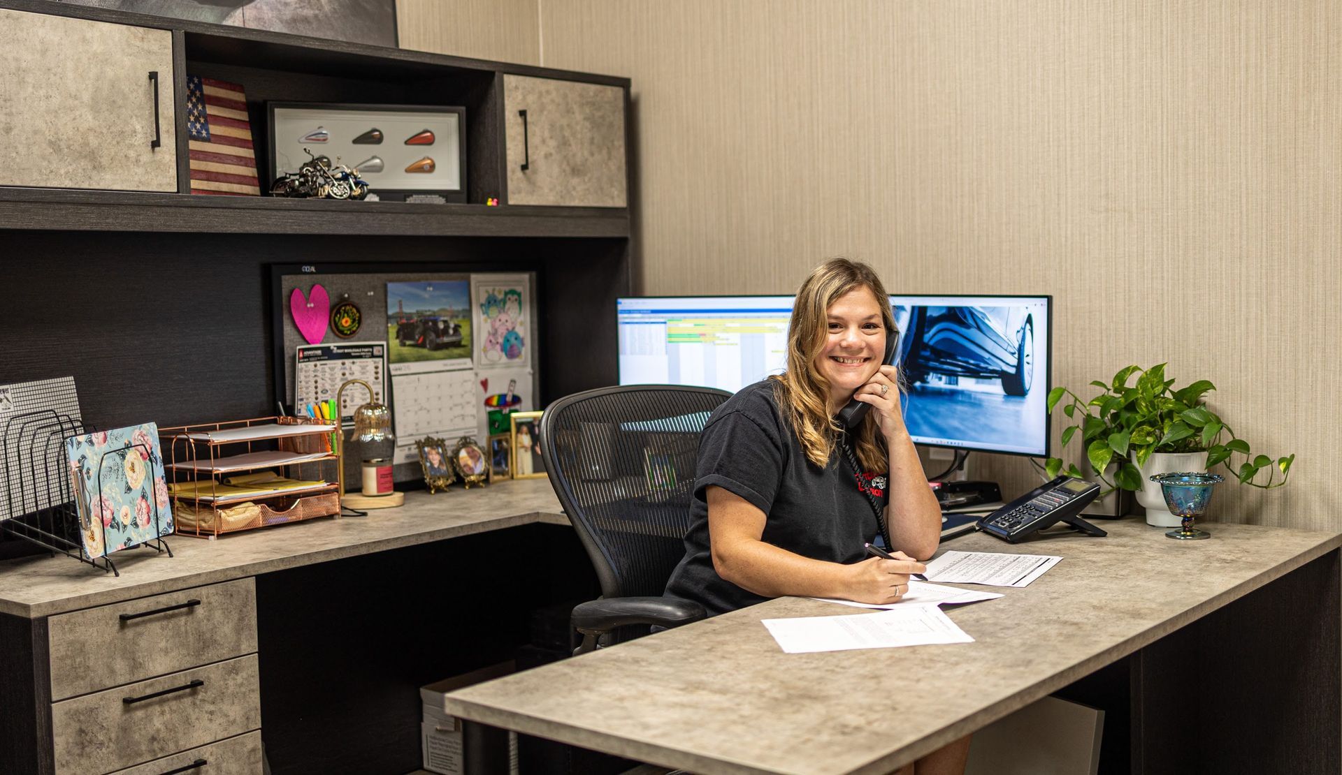 Woman seated at a desk in a modern office, smiling beside a computer and papers, with plants and shelves nearby