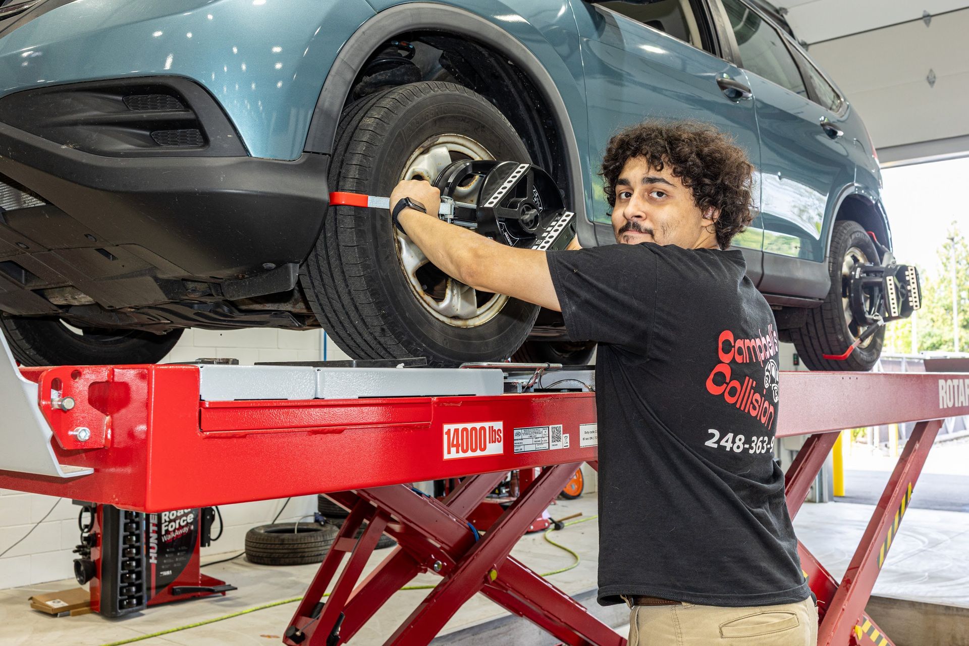 Mechanic working under a teal car on a red lift in an auto repair shop