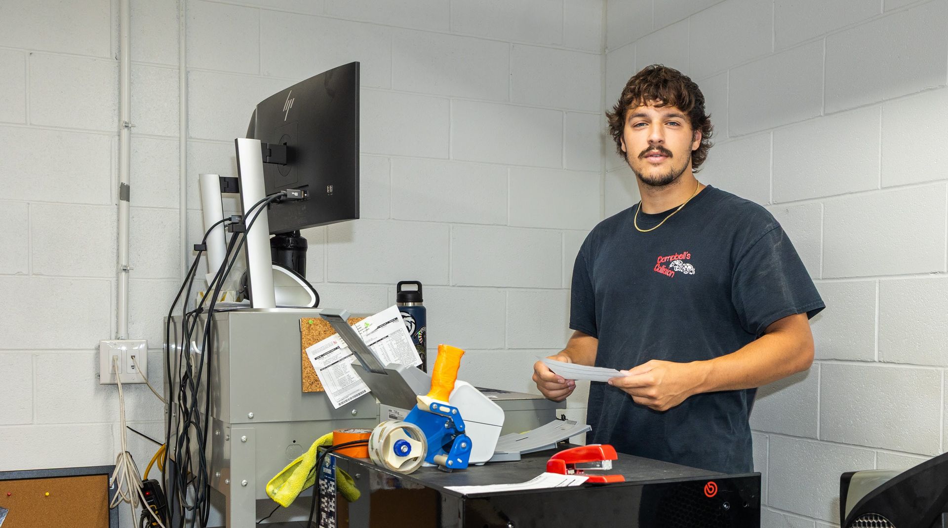 Person standing in a workshop beside a cluttered workbench and computer monitor, holding papers.