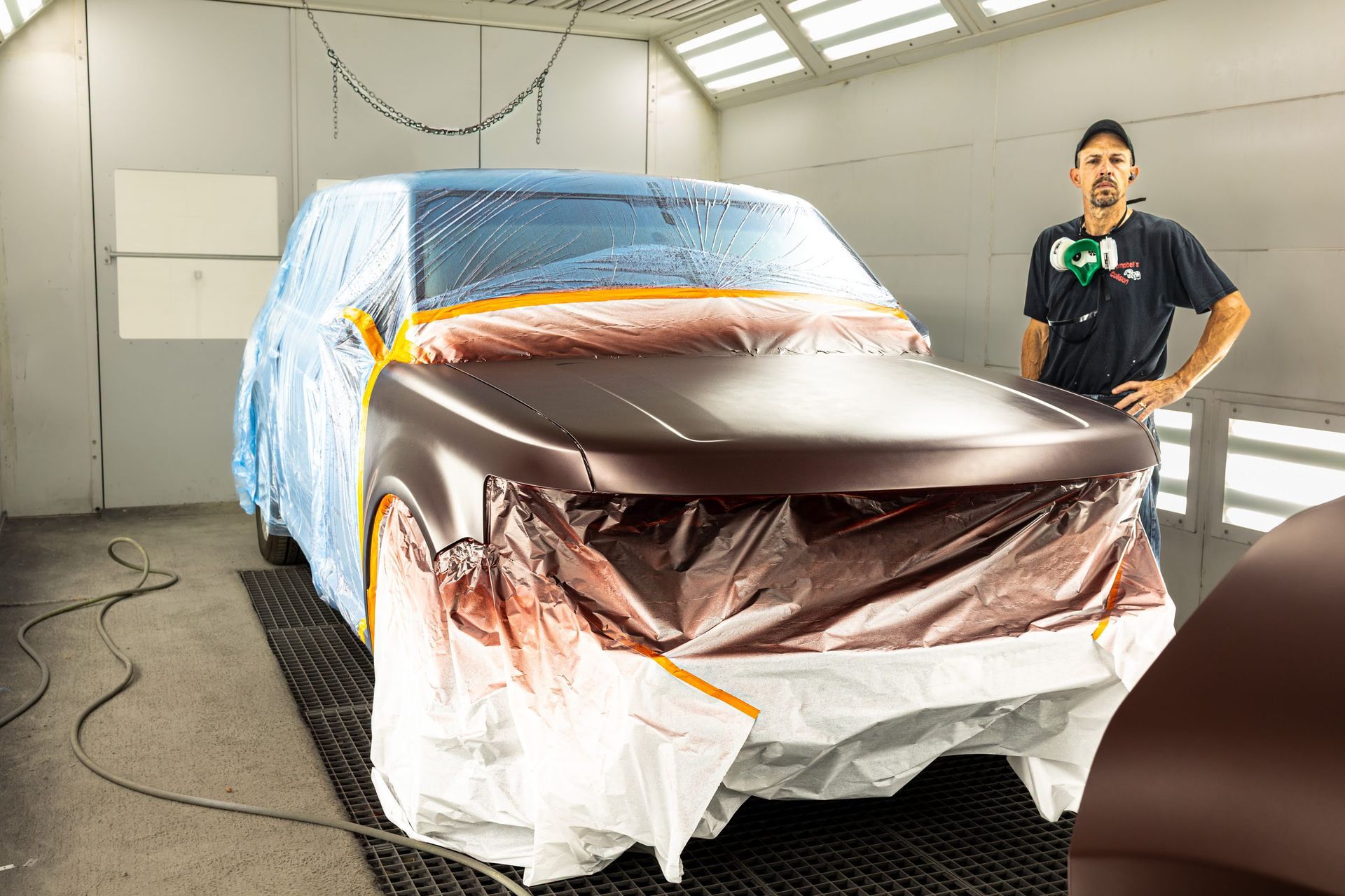 Masked car in spray booth, covered with plastic and paper, with a worker standing beside it.