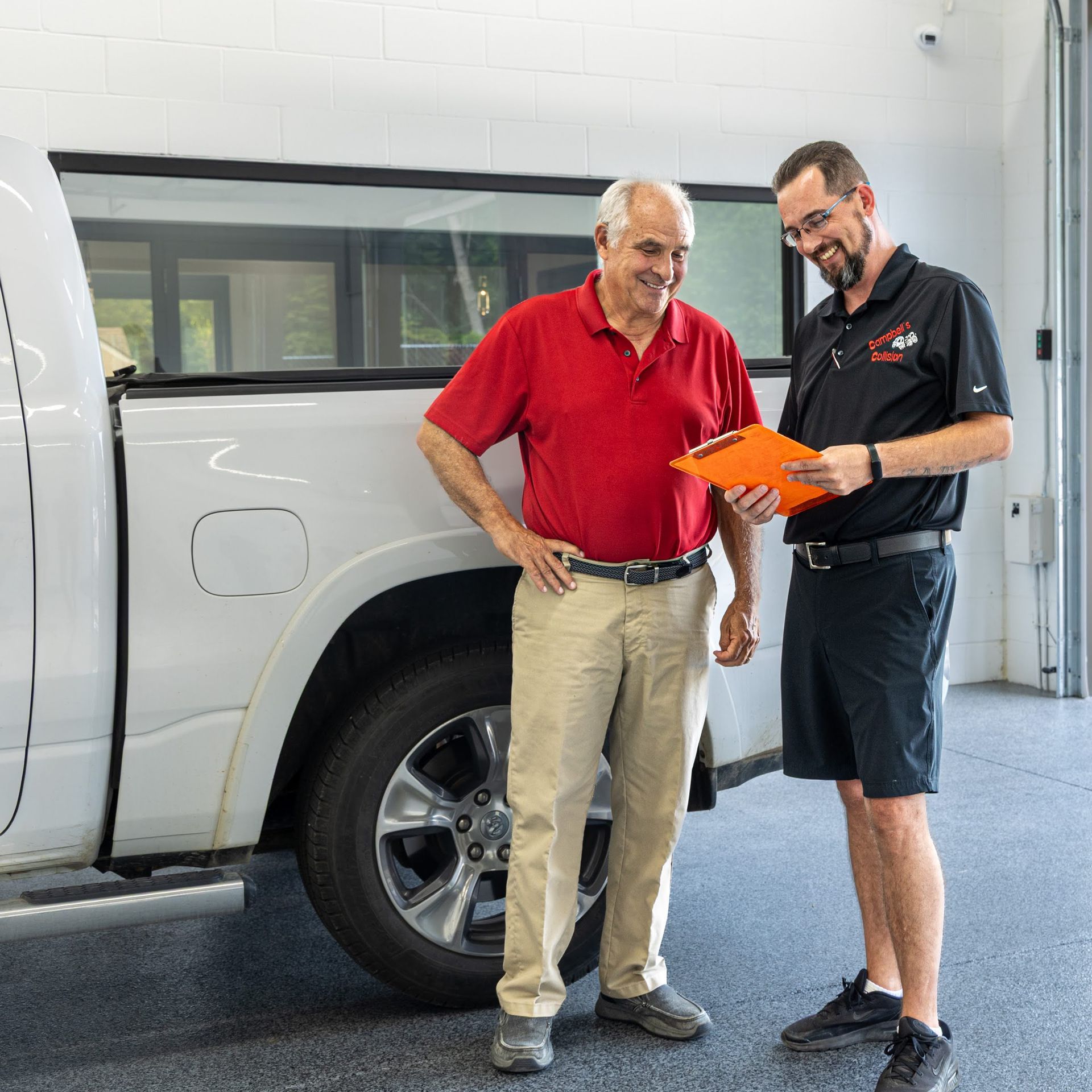 Two men talking beside a white pickup truck in a service bay, one holding an orange clipboard