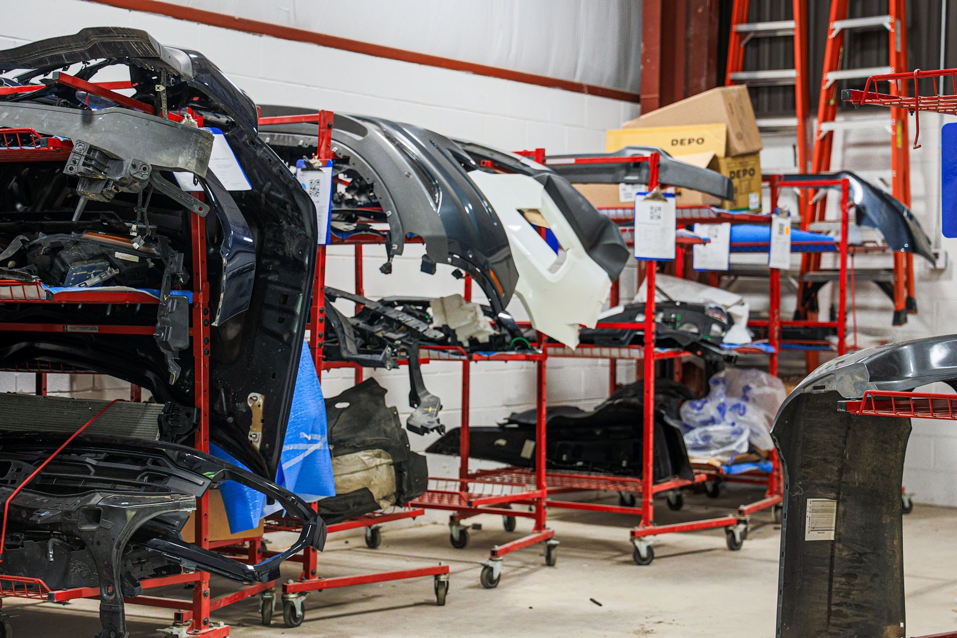 Automotive parts stacked on red metal racks in a workshop or warehouse, with car body panels in black and white.