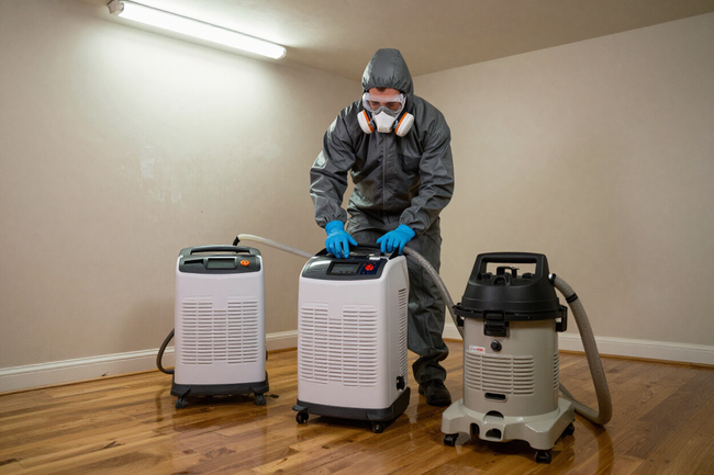 Worker in mask and gloves operating two white dehumidifiers in a basement-like room with wood floor
