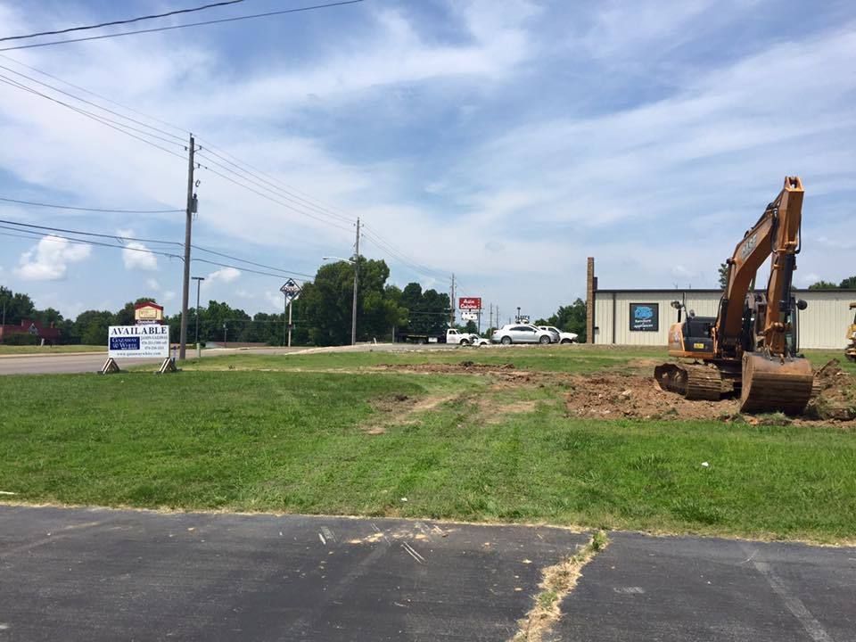 Construction excavator on grassy roadside near a small building and parked vehicles under a cloudy sky