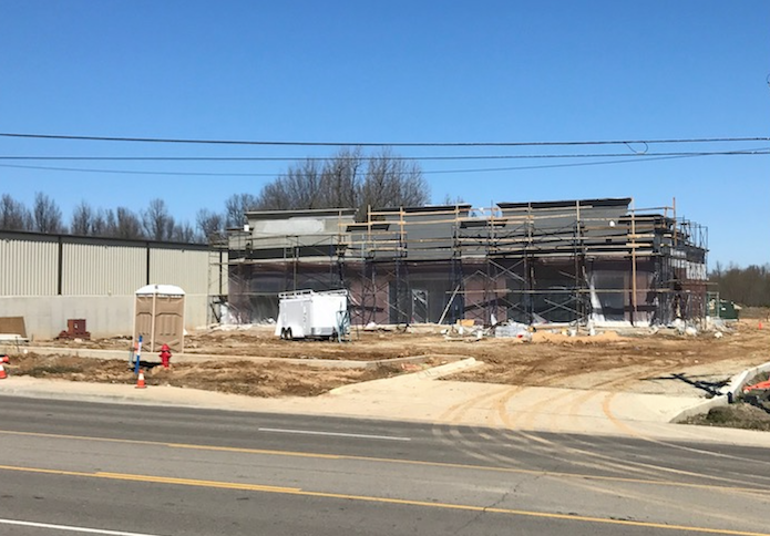 Construction site with partially built steel-frame building, dirt lot, and road in foreground under blue sky