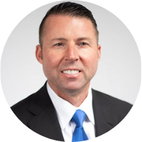 Professional headshot of a man in a dark suit, white shirt, and blue tie against a light gray background