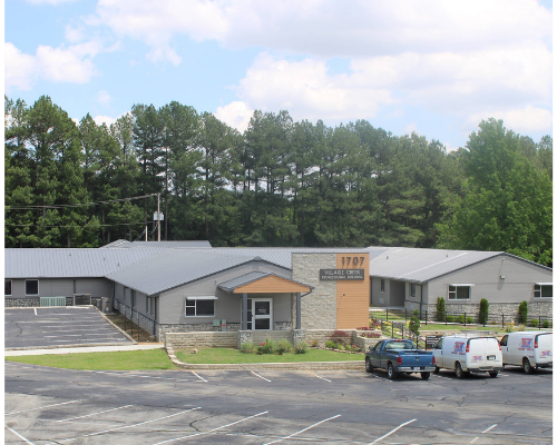 Small building with parking lot and trees in the background under a cloudy sky