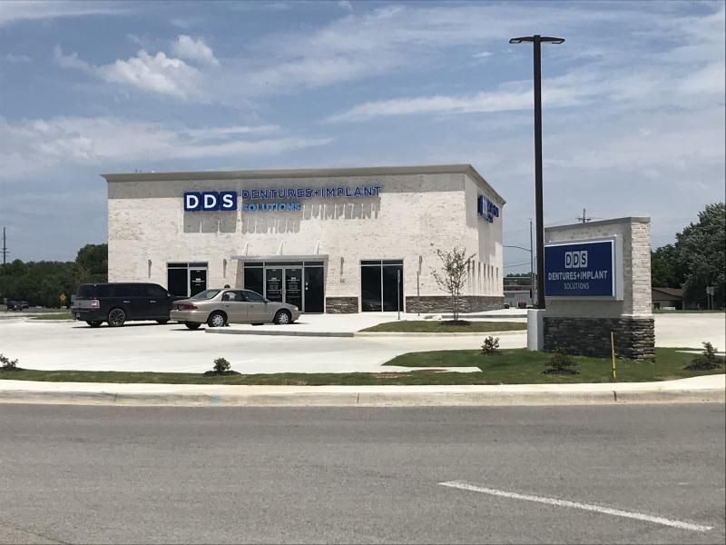 Small DDS dental office with white stone exterior, blue signs, parked cars, and roadside entrance