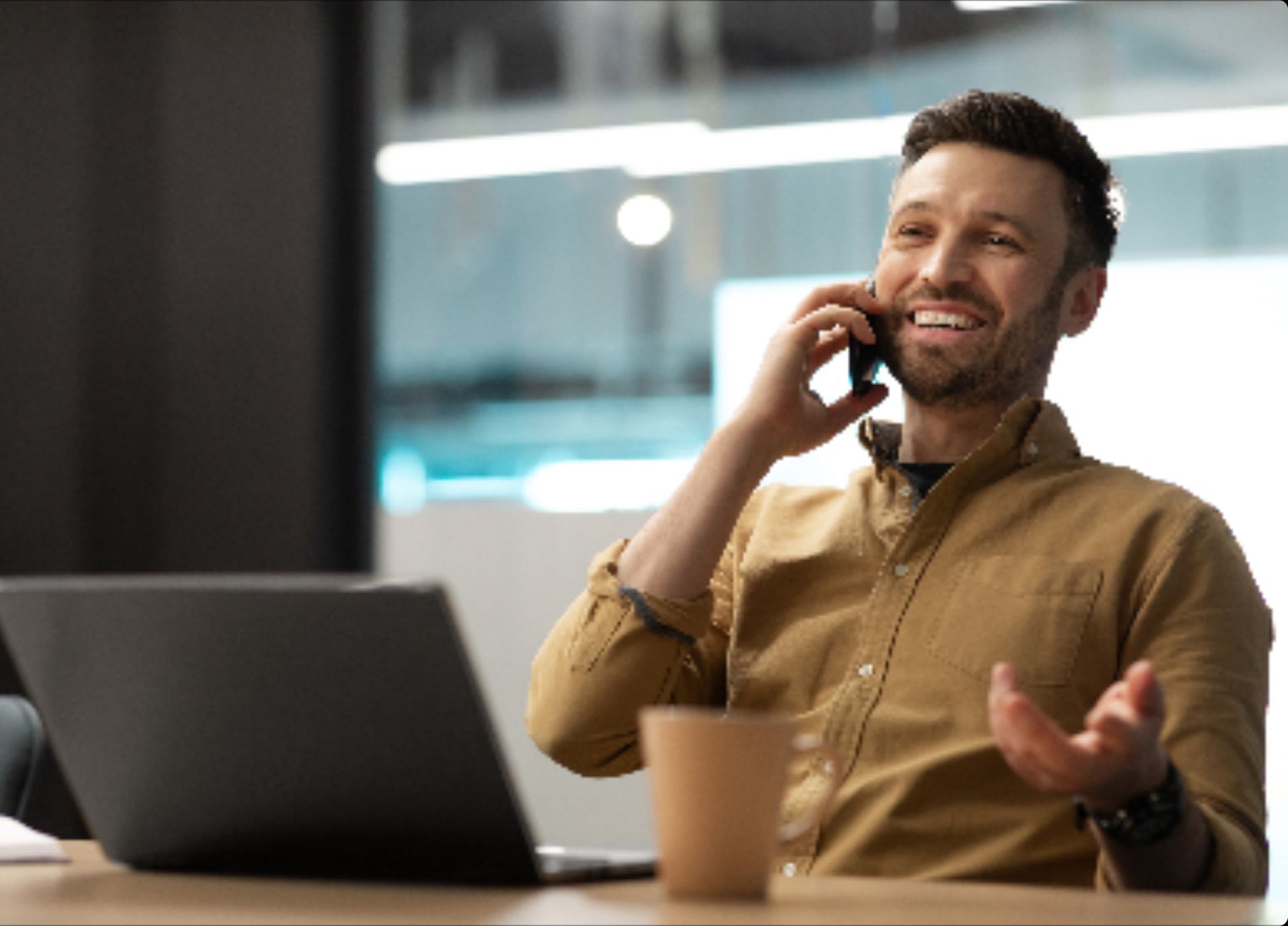 A person in a tan shirt smiles while talking on a smartphone at a desk with a laptop and a cup.