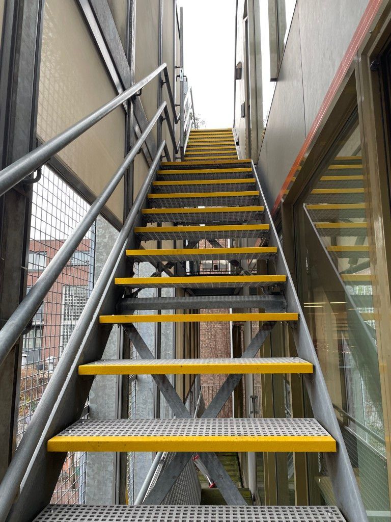 A set of metal stairs with yellow railings leads to a small platform on a grassy embankment next to a water channel.
