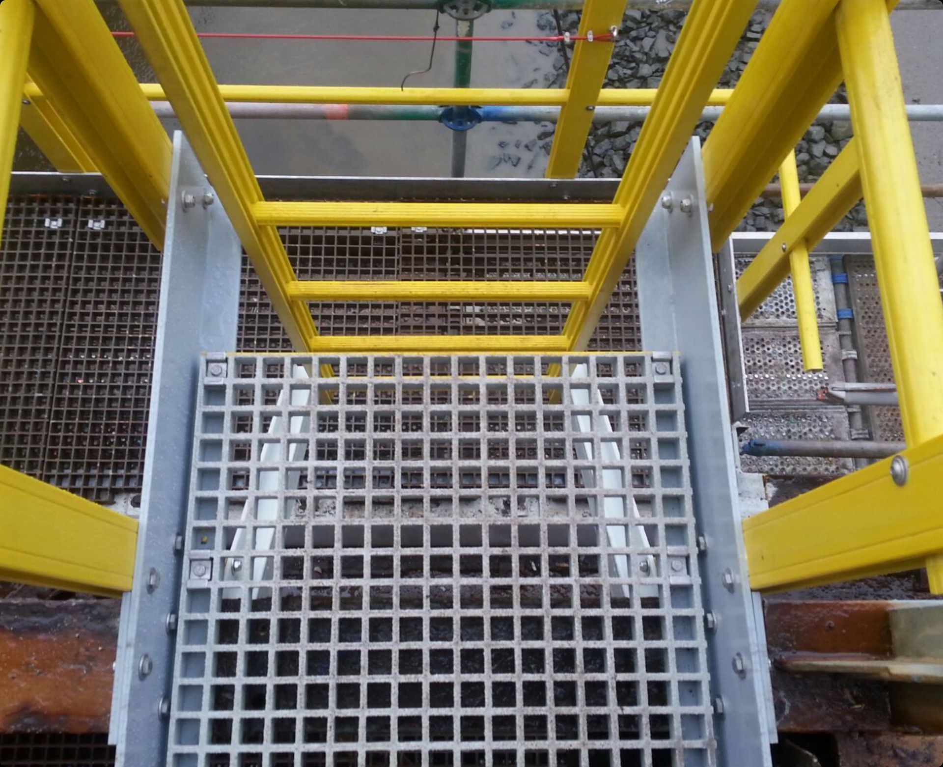 A top-down view of metal grating stairs with yellow railings leading to a work platform above water.