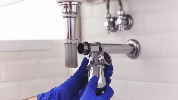 Blue-gloved hands adjust a chrome sink drain pipe under a white bathroom sink.