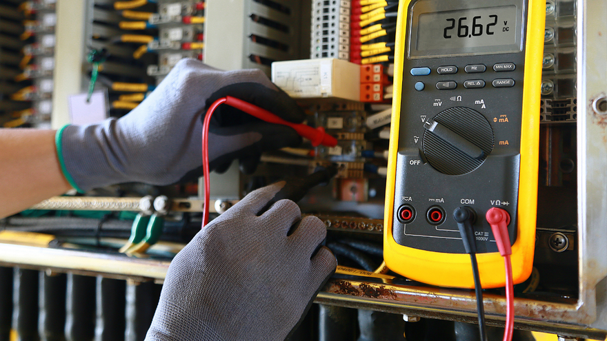 Gloved hands testing electrical wiring with a yellow digital multimeter in a workshop