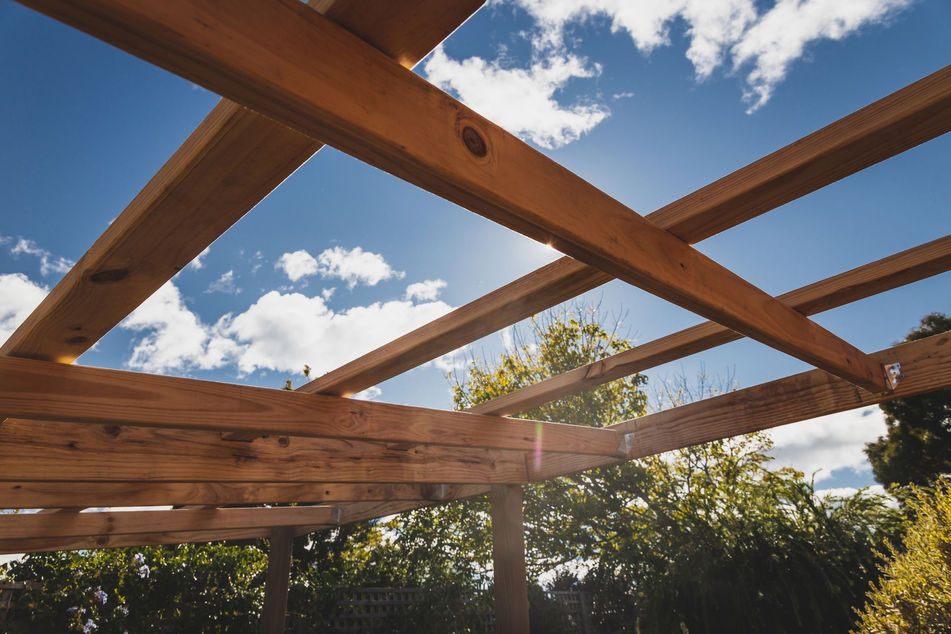 Wooden pergola beams framing a blue sky with white clouds above trees