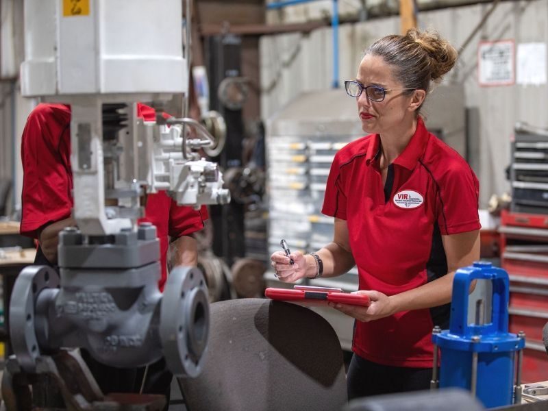 Worker in red uniform holds a clipboard beside industrial machinery in a factory.