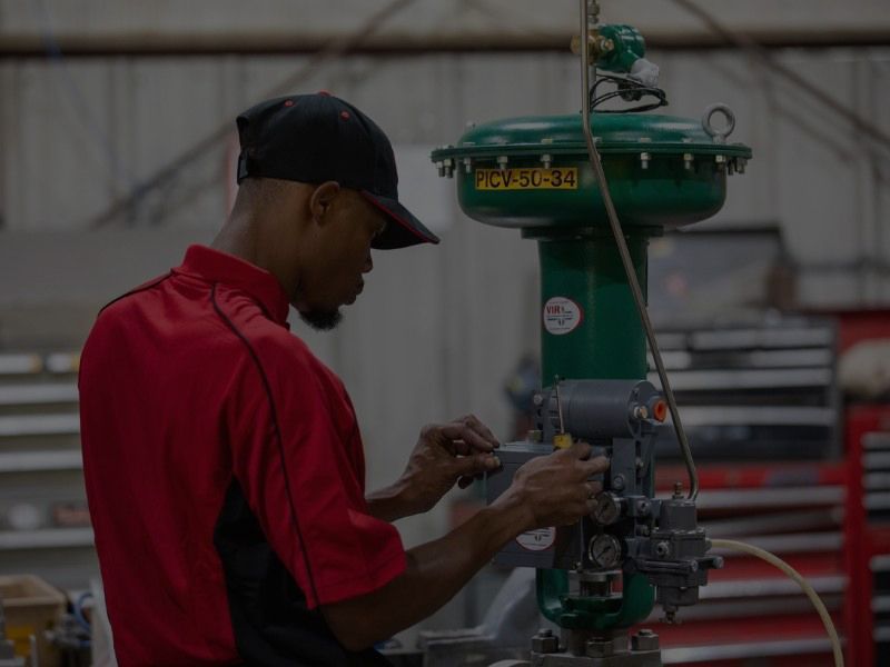Technician in a red shirt and black cap works on green industrial equipment in a workshop.