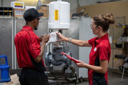 Two workers in red shirts talking in a factory beside machinery and clipboards
