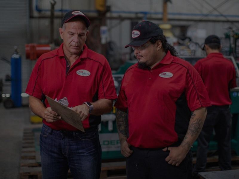 Two workers in red uniforms discussing a clipboard in an industrial warehouse
