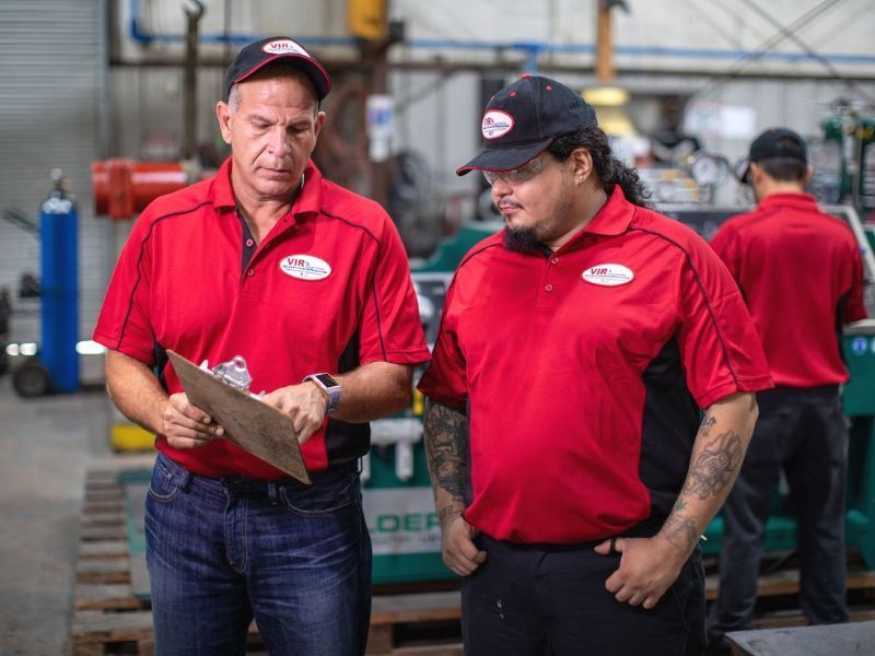 Two workers in red shirts review a clipboard in a warehouse setting.