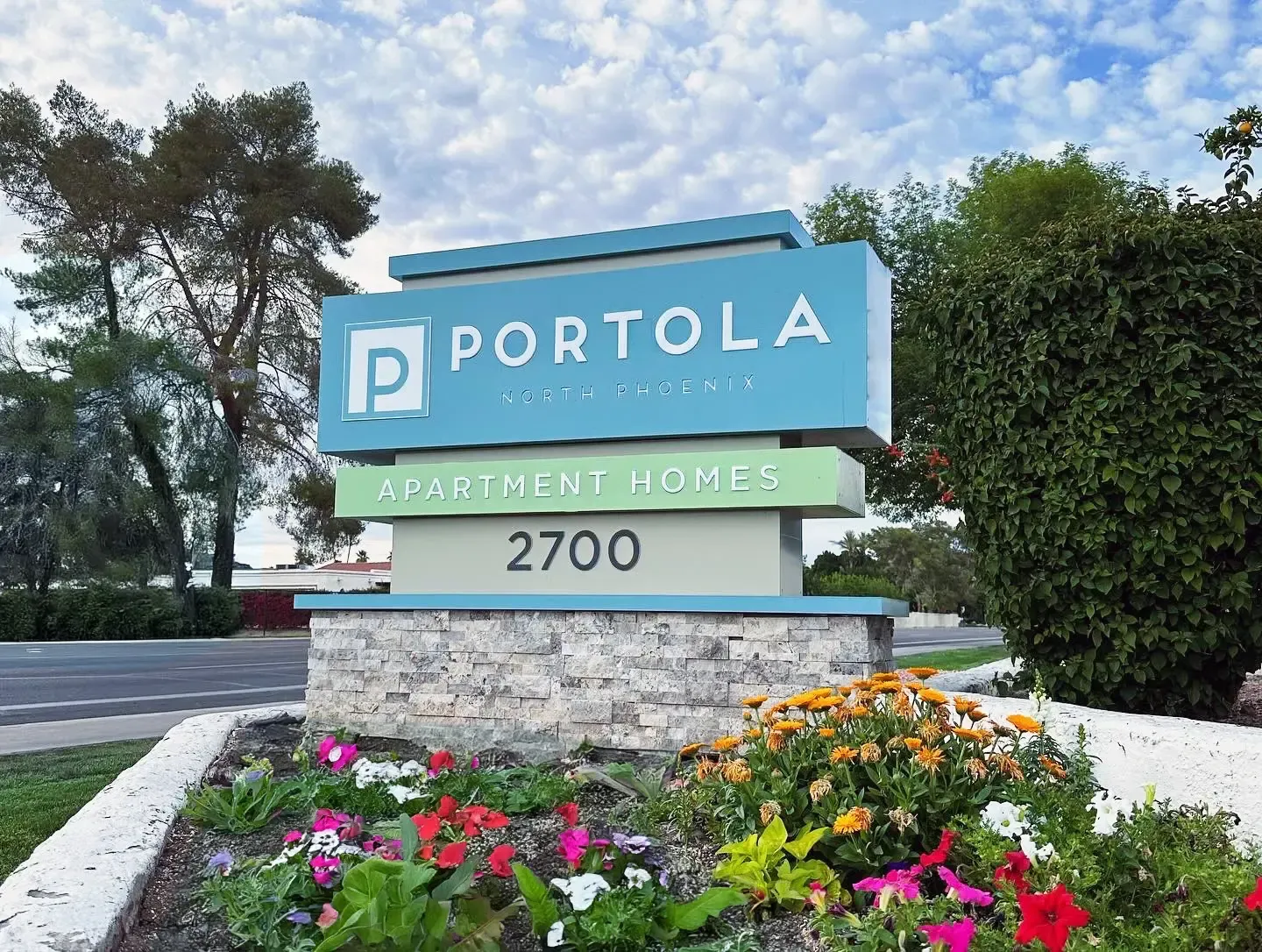 A sign for portola apartments is surrounded by flowers and trees.