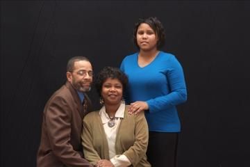 Three people posing against a dark backdrop, one seated and two standing behind her.