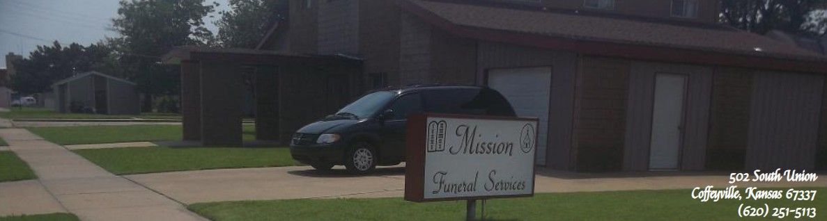 Residential street with a parked black car and a yard sign in front of a brick building.